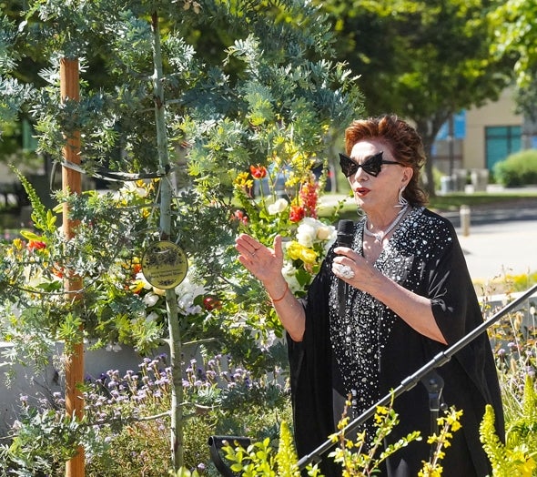 Maria Manetti Shrem dressed in a black dress with silver sparkles, wearing a statement necklace and ring, and bedazzled cat eye sunglasses stands at the tree planting dedication at UC Davis campus.