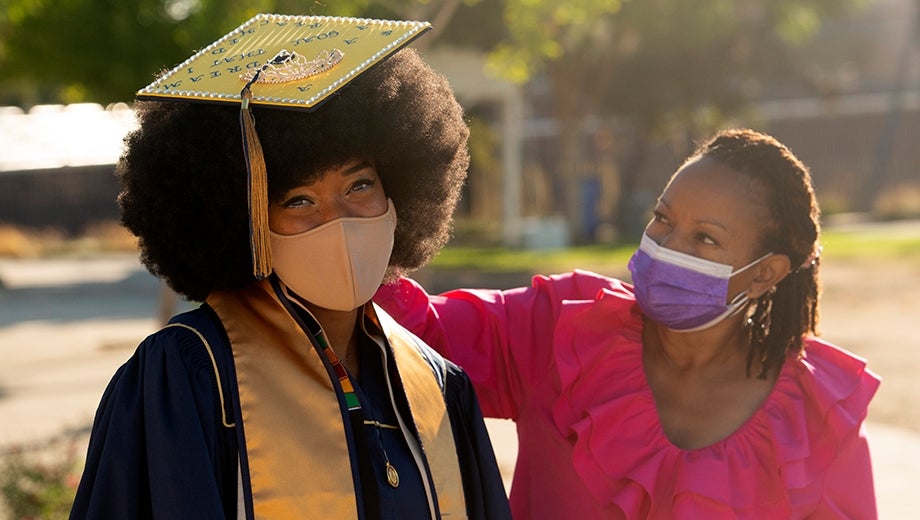 a masked woman graduate in a cap and gown smiling with a family member