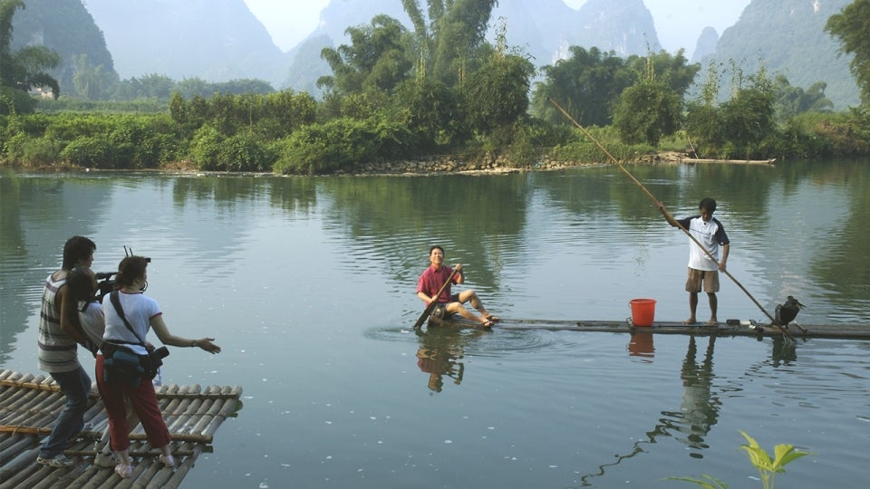 A crew films Martin Yan on location in Yangshuo, China. He is sitting in a long and skinny boat in a lake-like body of water.
