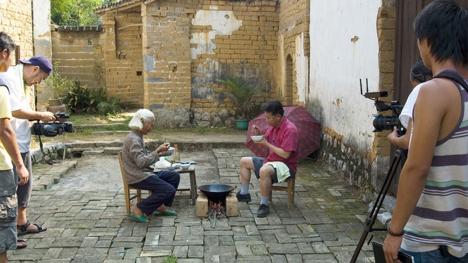 Martin Yan films a show in a village in Yangshuo, China. He is sitting in a small wooden chair across from an older lady with a fire between the two of them in a cobblestone alley.