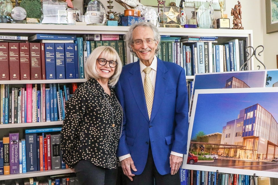 Anne-Marie and Dr. Schwab stand near bookshelves and a rendering of the new Tschannen Eye Center at the UC Davis Eye Center library.