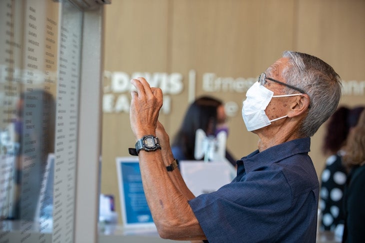 A man wearing a mask takes a photo with his phone of glass display containing names. 