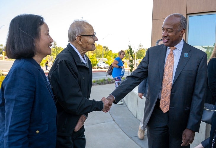 A woman in a blue jacket stands next to an elderly man who is shaking hands with a man in a blue suit.