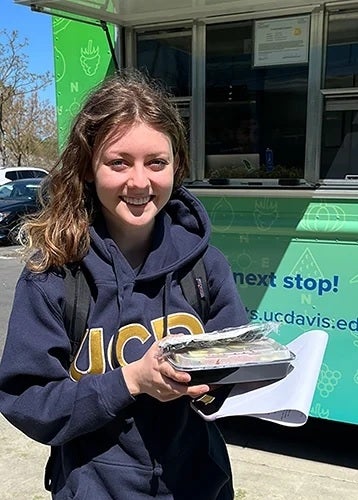 Photo of Alexandra Sarimsakci holding a tray of prepared food in front of the AggieEats food truck on campus.