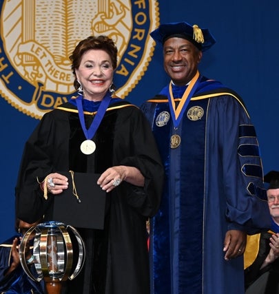 Maria Manetti Shrem wears her university medal in commencement regalia while holding onto her black grad cap and stands next to Chancellor Gary May in blue and gold faculty regalia.