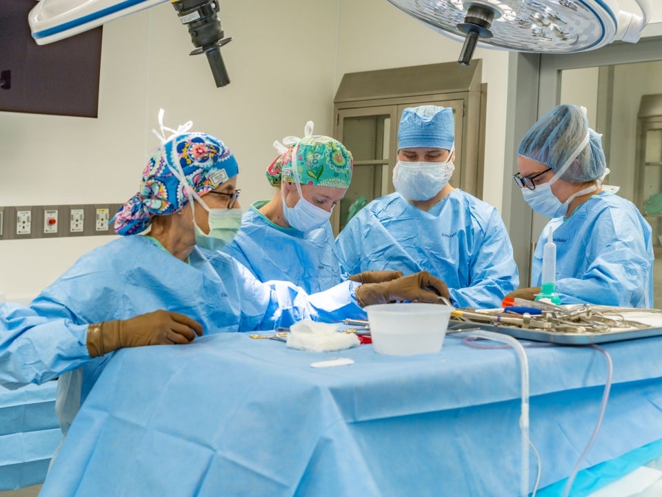 Four surgeons wearing blue scrubs and PPE look down on an operating table.