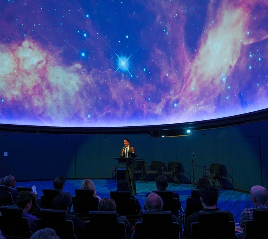 A presenter at an auditorium with a projection of the night sky with stars and planets.