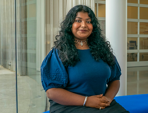 Madeline Furtado, a UC Davis undergraduate student seated with hands clasped, wearing a turquoise top in front of a large glass window in the Manetti Shrem Museum.