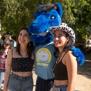 Gunrock the mascot wearing a light blue Give Day shirt poses with two students.