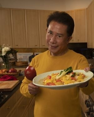 Chef Martin Yan in a mustard-colored sweater lifts up a plate of stir-fried vegetables and a red ladle.