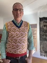 A portrait of artist Josh Faught wearing glasses, a green-and-white striped button-up shirt, and a patterned sweater vest with floral motifs, standing indoors in front of hanging textile artwork.