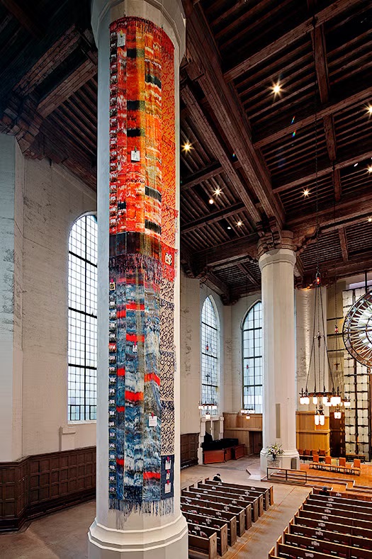 A long, colorful woven textile banner wrapped vertically around a tall white column inside a cathedral, transitioning from warm orange and red tones at the top to cool blues and grays at the bottom.