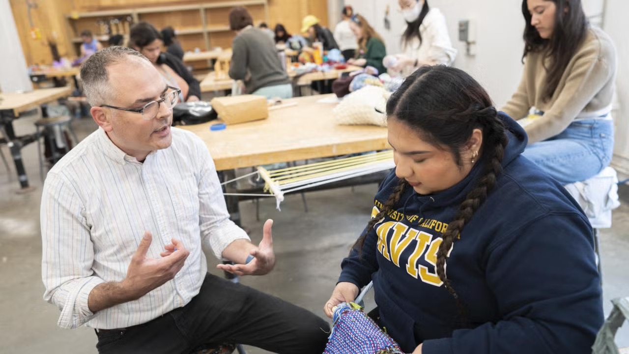 Artist Josh Faught, wearing glasses and a striped button-down shirt, gestures while speaking with a student in a UC Davis sweatshirt who is working on a hand-woven textile project in a classroom setting with other students in the background.