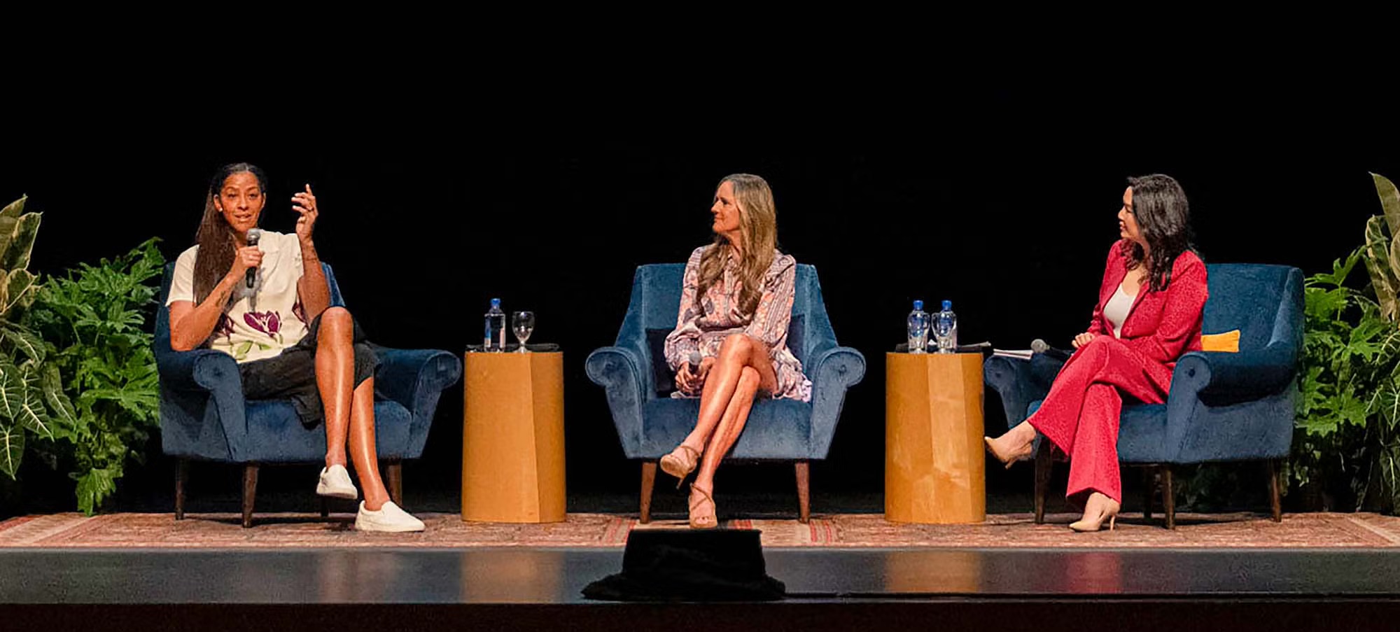 Three women on a dark stage panel, center woman gesturing while others listen