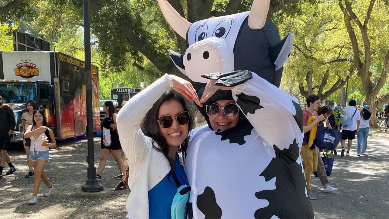 Two people wearing sunglasses pose while forming the shape of a heart on the Quad. One is wearing an inflatable cow costume.