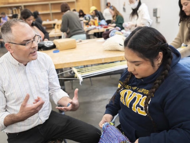Artist Josh Faught, wearing glasses and a striped button-down shirt, gestures while speaking with a student in a UC Davis sweatshirt who is working on a hand-woven textile project in a classroom setting with other students in the background.