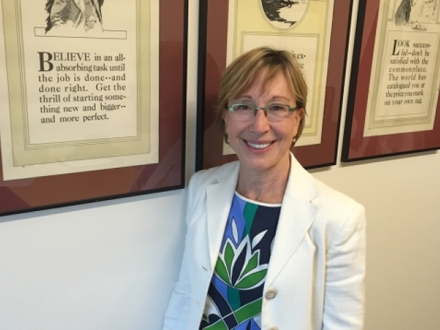 Hollis wearing a blue and green blouse with a white blazer and glasses next to a wall with framed pictures.