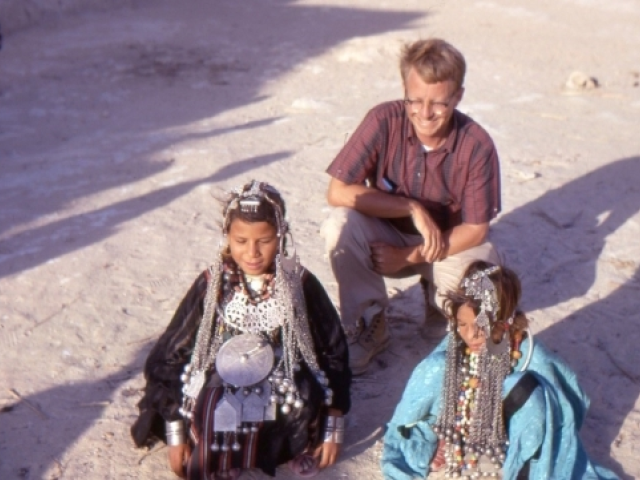 Louis crouches outdoors with two people wearing traditional garb with ornate jewelry and headpieces.