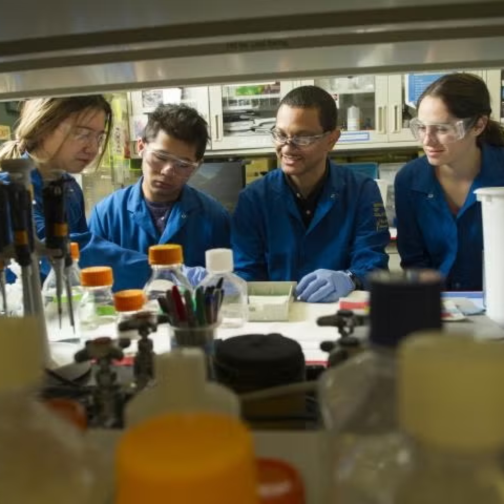 A group of scientists wearing blue lab coats and eye protection in a wet lab.