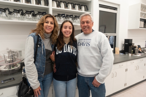 Two parents and their student pose in the UC Davis Coffee Center with a work station and shelf with coffee pots visible behind.
