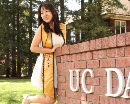 A graduate wearing a white dress and a gold grad stole paired with a blue and gold philanthropy cord poses next to the red brick UC Davis sign.
