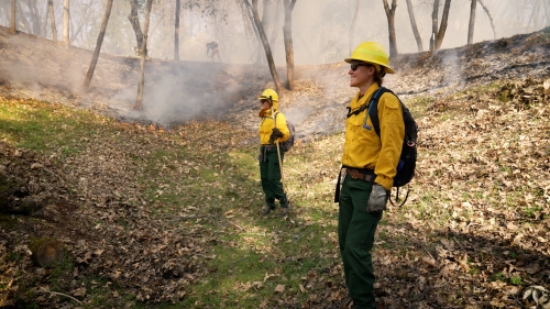 Two women in yellow firefighting in shirts and helmets stand in meadow with light smoke in background during prescribed burn.