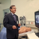Tom Nesbitt appearing to speak with someone via a computer screen in a health care setting.